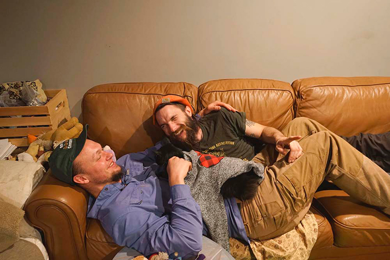 Two men share a laugh as they lay together on a couch with their cat.