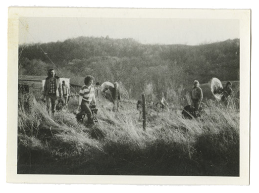 A black and white vintage photo scan shows women carrying tools in a grass feild.
